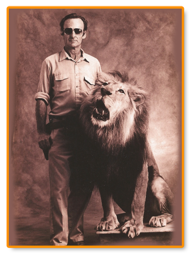 A sepia-toned image of Hubert Geza Wells standing next to a roaring lion. Wells is wearing sunglasses and a button-down shirt with rolled-up sleeves. The lion is seated beside him, baring its teeth. The image is framed with an orange border.